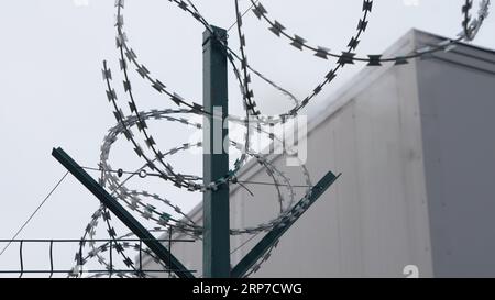 Stacheldraht auf der Oberseite des Zauns vor dem Hintergrund eines Teils des grauen Gebäudes Stockfoto