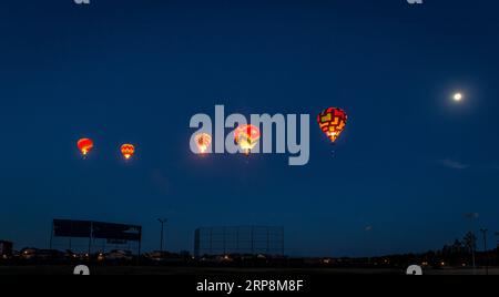 Fünf Heißluftballons fliegen bei Tagesanbruch über ein Baseballfeld. Alle fünf Ballons werden von ihren Gasdüsen angezündet. Der Himmel ist ein dunkles Blau, das man auf der blu sieht Stockfoto