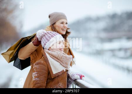 Glückliche stilvolle Mutter und Kind in Mantel, Hut, Schal und Fäustlingen mit Einkaufstaschen im Winter in der Stadt. Stockfoto