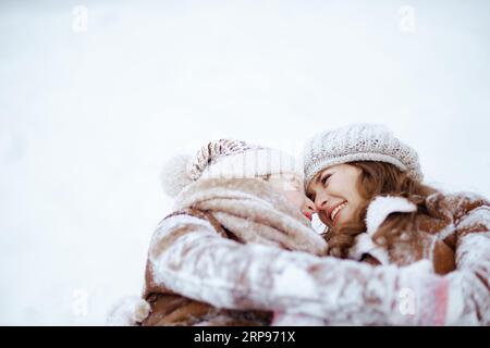 Lächelnde, stilvolle Mutter und Tochter in Mantel, Hut, Schal und Fäustlingen liegen im Winter im Schnee im Stadtpark. Stockfoto