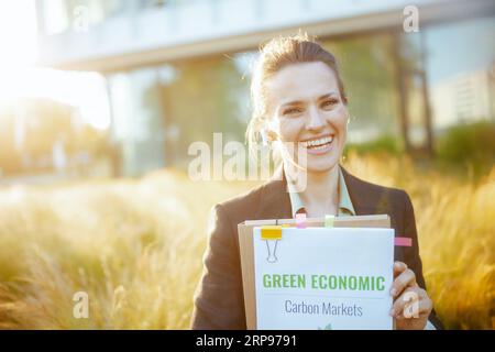 Lächelnde, moderne Angestellte in schwarzer Jacke in der Nähe des Business Centers mit Dokumenten, grünen Sparplänen, Ordnern und kabellosen Kopfhörern. Stockfoto