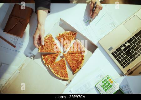 Nachhaltiger Arbeitsplatz. Draufsicht auf weibliche Mitarbeiterin im grünen Büro mit Pizza und Laptop. Stockfoto