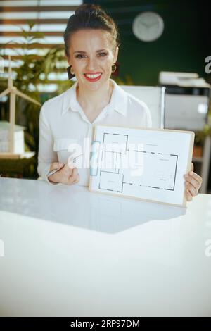 Zeit, weiterzumachen. Lächelnde elegante 40-jährige Immobilienmaklerin im modernen grünen Büro in weißer Bluse mit Klemmbrett und Bauplan. Stockfoto