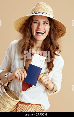 Strandurlaub. Lächelnde moderne Frau mittleren Alters in weißer Bluse und Shorts isoliert auf beigem Hintergrund mit Strohsack, Flugticket und Strohhut. Stockfoto