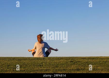 Frau, die im Lotus sitzt, posiert auf grünem Gras über blauem Himmel, verbindet sich mit dem Naturkonzept Stockfoto