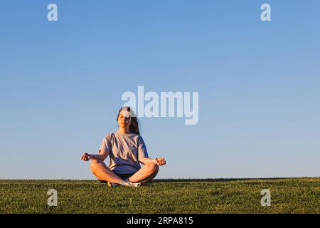 Frau sitzt in einer Meditationsposition auf dem grünen Rasen über dem blauen Himmel, Achtsamkeitskonzept Stockfoto