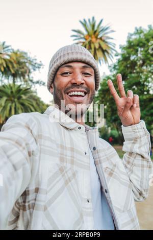 Vertikales Einzelporträt eines jungen gutaussehenden Mannes, der lächelnd ein Selfie-Portrait mit einem Smartphone macht, das das Friedenszeichen mit den Fingern in einem Park im Freien mit einem Beanie-Hut macht. Lifestyle-Konzept. Hochwertige Fotos Stockfoto
