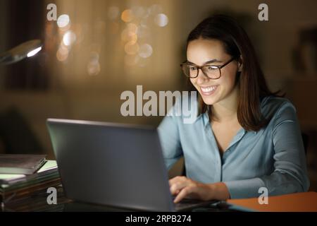 Glücklicher Telearbeiter mit Brille, der nachts zu Hause einen Laptop benutzt Stockfoto