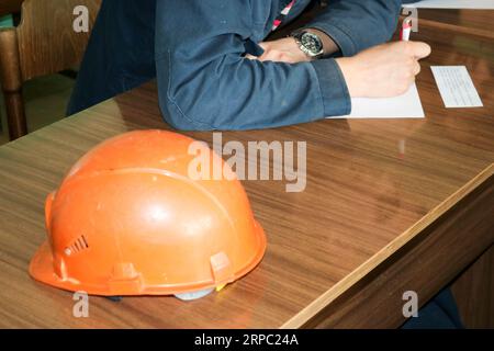 Ein Mann, der als Ingenieur mit einem orangegelben Helm auf dem Tisch arbeitet, studiert und schreibt in einem Notebook in einer Fabrik in einem Industriewerk. Stockfoto