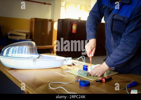 Ein Mann, der Elektriker arbeitet, sammelt den Stromkreis einer großen weißen Straßenlaterne mit Drähten, ein Relais in einer Fabrik eines Industriewerkes. Stockfoto