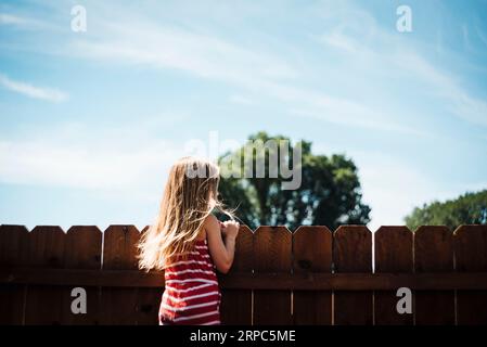 Das Mädchen schaut an einem sonnigen Sommertag über den Zaun im Hinterhof Stockfoto