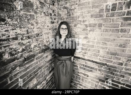 Eine junge Frau mit Brille und Kleid steht vor einer Ziegelmauer Stockfoto