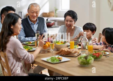 Junge asiatische Frau, die Bilder mit der Familie der 3. Generation auf dem Handy teilt, während sie zusammen isst Stockfoto