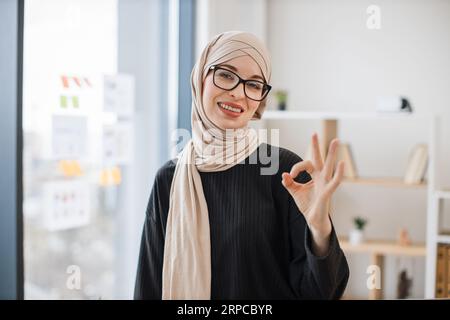 Porträt eines lächelnden arabischen Erwachsenen in Brillen, der auf einem Holzschreibtisch sitzt und die Hände auf der Brust gefaltet hat, mit dem Zeichen OK Stockfoto