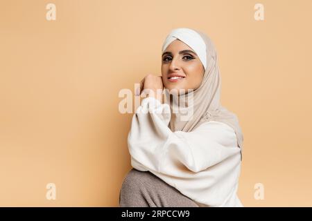 Moderne muslimische Frau mit Make-up in Hijab und weißem Sweatshirt, während sie auf Beige vor der Kamera lächelt Stockfoto