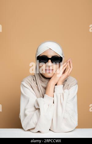 Unabhängige muslimische Frau in Hijab, Sonnenbrille und weißem Sweatshirt, die am Tisch auf Beige sitzt Stockfoto