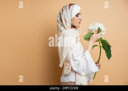 Anmutige muslimische Frau in Seidenkopftuch und Bluse mit weißer Blume auf Beige, Seitenansicht Stockfoto