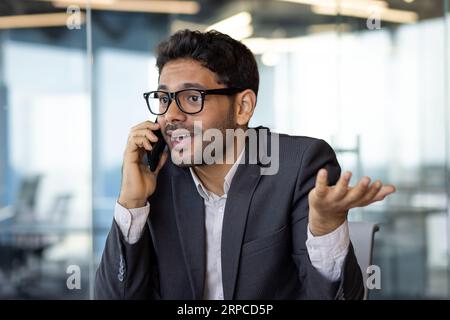 Unzufriedener und verärgerter Mann, der am Telefon aus nächster Nähe sprach, ein junger Geschäftsmann in Geschäftskleidung am Arbeitsplatz im Büro. Stockfoto