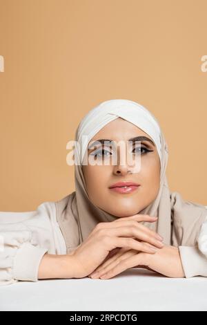 Positive und stilvolle muslimische Frau mit Make-up sitzt am weißen Tisch im Hijab und lächelt vor der Kamera Stockfoto
