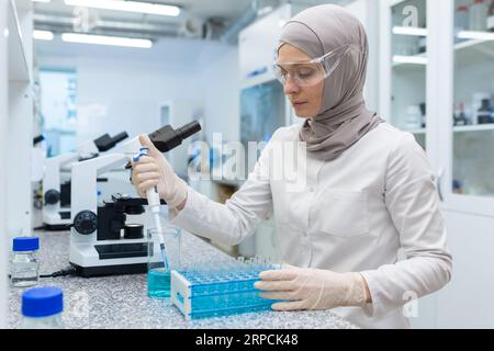 Muslimische junge Frau im Hijab, Wissenschaftlerin und Apothekerin im Labor. Sitzen am Tisch und Arbeiten mit Flüssigkeiten und Kolben. Stockfoto