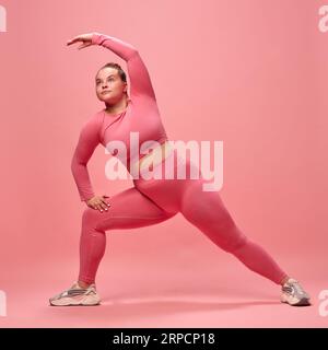 Ganzkörperporträt einer jungen Chubbu-Sportlerin im rosa Oberteil und Legging-Training, Stretching der Beine und Hände isolierter Studiohintergrund. Stockfoto