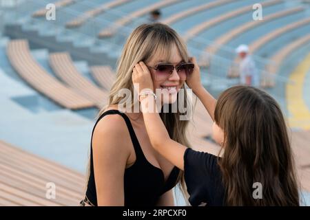 Das kleine Mädchen hilft der Mutter, am sonnigen Sommerabend beim Spaziergang im Park eine Sonnenbrille anzuziehen. Glückliche junge Familie, die Spaß hat und Zeit miteinander verbringt Stockfoto