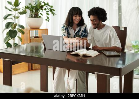 Glückliche junge Frau, die sich die Anzahl der von ihrem Freund gehaltenen Kreditkarten anschaut, während sie neben ihm sitzt und Bankdaten eingibt Stockfoto