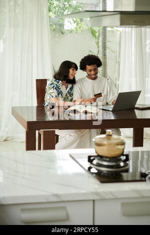 Glücklicher junger Mann, der sich die Kreditkarte anschaut und seine Nummer auf der Website eingibt, während er mit seiner Frau zu Hause Online-Waren bestellt Stockfoto