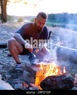 Ein brutaler Mann mit Bart kocht Essen in einer Bratpfanne auf einem Feuer in der Natur. Am Flussufer wird Speck gekocht. Camping, Sommer Stockfoto