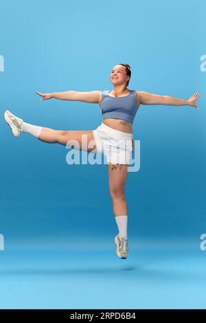 Foto in voller Länge von lächelnder übergewichtiger Frau, die Sportbekleidung trägt und Übungen auf blauem Studiohintergrund macht. Stockfoto