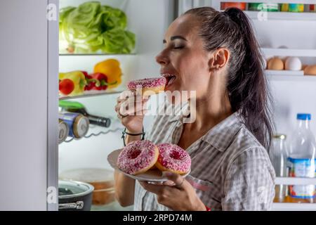 Hungrige Brünette im Pyjama genießt süße Donuts spät in der Nacht am offenen Kühlschrank. Stockfoto