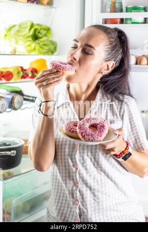 Hungrige Brünette im Pyjama genießt süße Donuts spät in der Nacht am offenen Kühlschrank. Stockfoto