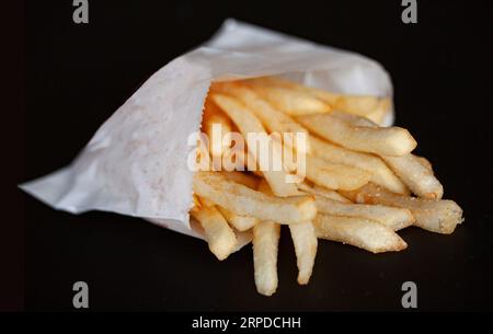 Frische Pommes frites Pommes frites Pommes frites Pommes frites in einem kleinen weißen Papierbeutel isoliert auf schwarzem Hintergrund. Selektiver Fokus mit geringer Schärfentiefe. Stockfoto