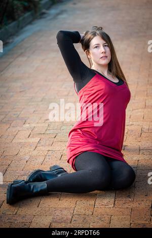 Eine schöne junge Frau, die ein rotes Kleid und schwarze Strümpfe trägt und auf einem gemauerten Pfad in einer legeren Pose sitzt Stockfoto