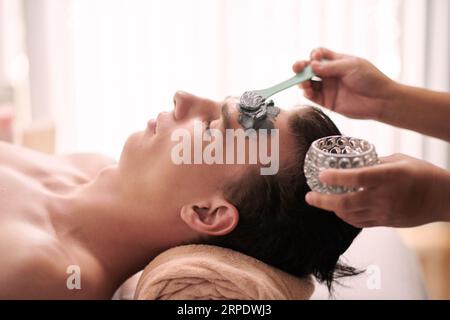 Seitenansicht eines jungen, ruhigen Mannes mit geschlossenen Augen, der den Kopf auf einem gerollten, weichen Handtuch hält, während eine Kosmetikerin eine Tonmaske auf sein Gesicht aufträgt Stockfoto
