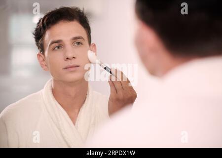 Junger Mann in einem weißen weichen Bademantel, der morgens vor dem Spiegel im Badezimmer steht und Rasierschaum mit Bürste auf das Gesicht aufträgt Stockfoto