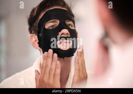 Junger Mann mit schwarzer Feuchtigkeitslaken-Maske auf dem Gesicht, der morgens vor dem Spiegel im Badezimmer steht und sich selbst anschaut Stockfoto
