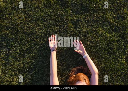 Nicht erkennbare Frau, die auf dem grünen Gras liegt, mit erhobenen Händen, Blick von oben Stockfoto