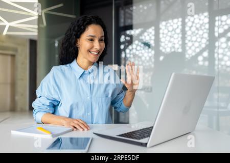 Junge erfolgreiche hispanofrau, die im Büro mit Laptop arbeitet, mit Videoanruf vor der Kamera winkt, Geschäftsfrau, die mit Kollegen online aus der Ferne spricht. Stockfoto