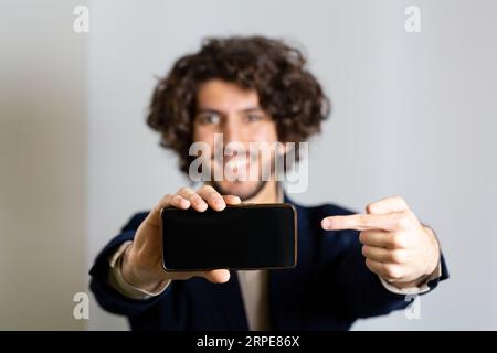 Junger lächelnder Mann mit Smartphone in der Hand, der mit dem Finger auf den schwarzen Bildschirm zeigt. Weißer Werbeträger. Selektiver Fokus auf Mobilgeräte und verschwommene BA Stockfoto