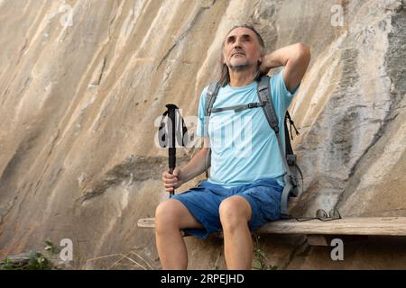 Ein grauhaariger erwachsener Mann mit Gehstöcken sitzt auf einer Holzbank unter einem Felsen und schaut nach oben Stockfoto