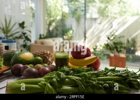 Vielfalt an frischem Obst und Gemüse in verschiedenen Farben, die auf dem Küchentisch vor der Kamera gegen die häusliche Umgebung und große Fenster liegen Stockfoto