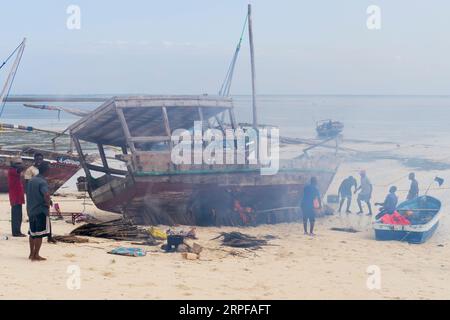 Afrikanische Fischer setzten Feuer unter ihrem traditionellen hölzernen Dhow-Boot am Strand, um es vor Schädlingen und Holzwürmern zu schützen und das Boot zu reparieren. Sansibar, Tansan Stockfoto