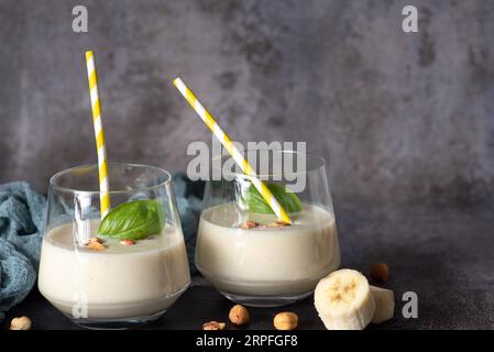 Frischer, gesunder Smoothie mit Banane und Haselnüssen, in einem Glasbecher mit gelb-weißem Trinkschlauch Stockfoto