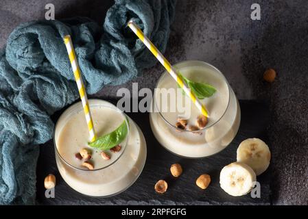Frischer, gesunder Smoothie mit Banane und Haselnüssen, in einem Glasbecher mit gelb-weißem Trinkschlauch Stockfoto