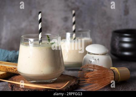 Frischer, gesunder Smoothie mit Banane und Birne, in einem Glasbecher mit schwarz-weißem Trinkschlauch, auf einem Holztablett und grauem Hintergrund Stockfoto