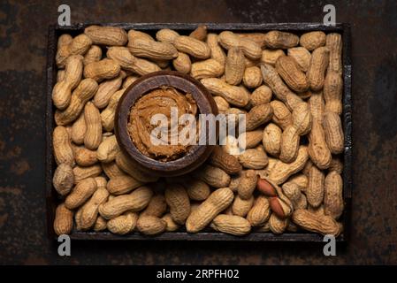 Cremige, frisch zubereitete Erdnussbutter in einem Holzglas und Haufen von geschälten Erdnüssen herum Stockfoto