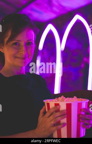 Glückliches Mädchen mit Popcorn-Box in Neonlichtern. Popcorn-Hintergrund im Kino. Junge Frau im Kino. Freizeitaktivitäten. Entertainment-Konzept. Stockfoto