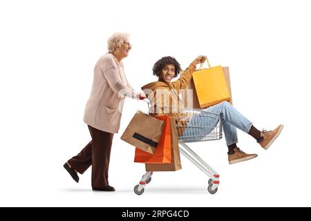 Großmutter schiebt einen jungen afroamerikanischen Mann mit Einkaufstaschen in einen Warenkorb isoliert auf weißem Hintergrund Stockfoto