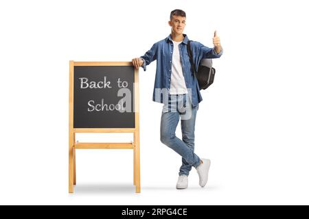 Portrait eines männlichen Teenagers mit einem Rucksack, der auf eine Tafel gestützt ist, mit Text zurück zur Schule und Daumen nach oben getippt, isoliert auf wh Stockfoto